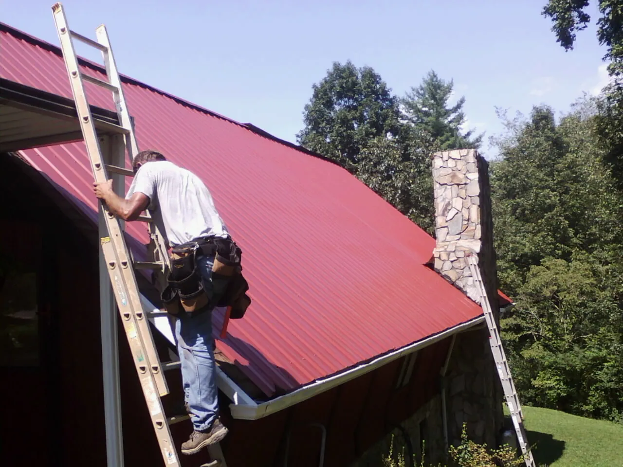 Roofer on a ladder beside a red metal roof and stone chimney — Soffit & Fascia Repair by Roofing Brothers in Glenn Dale